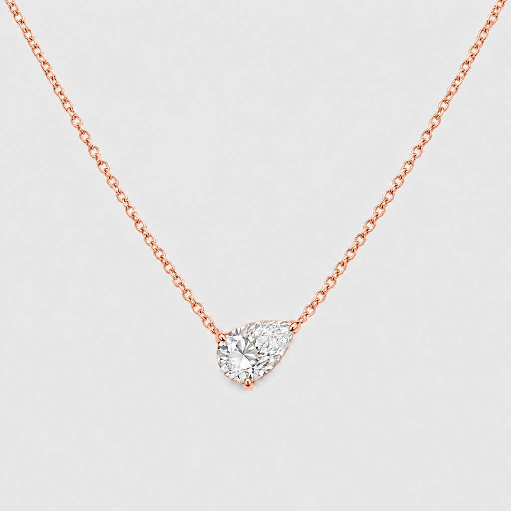 A rose gold-toned chain necklace with a pear-cut clear gemstone pendant in a prong setting, shown on a light gray background.