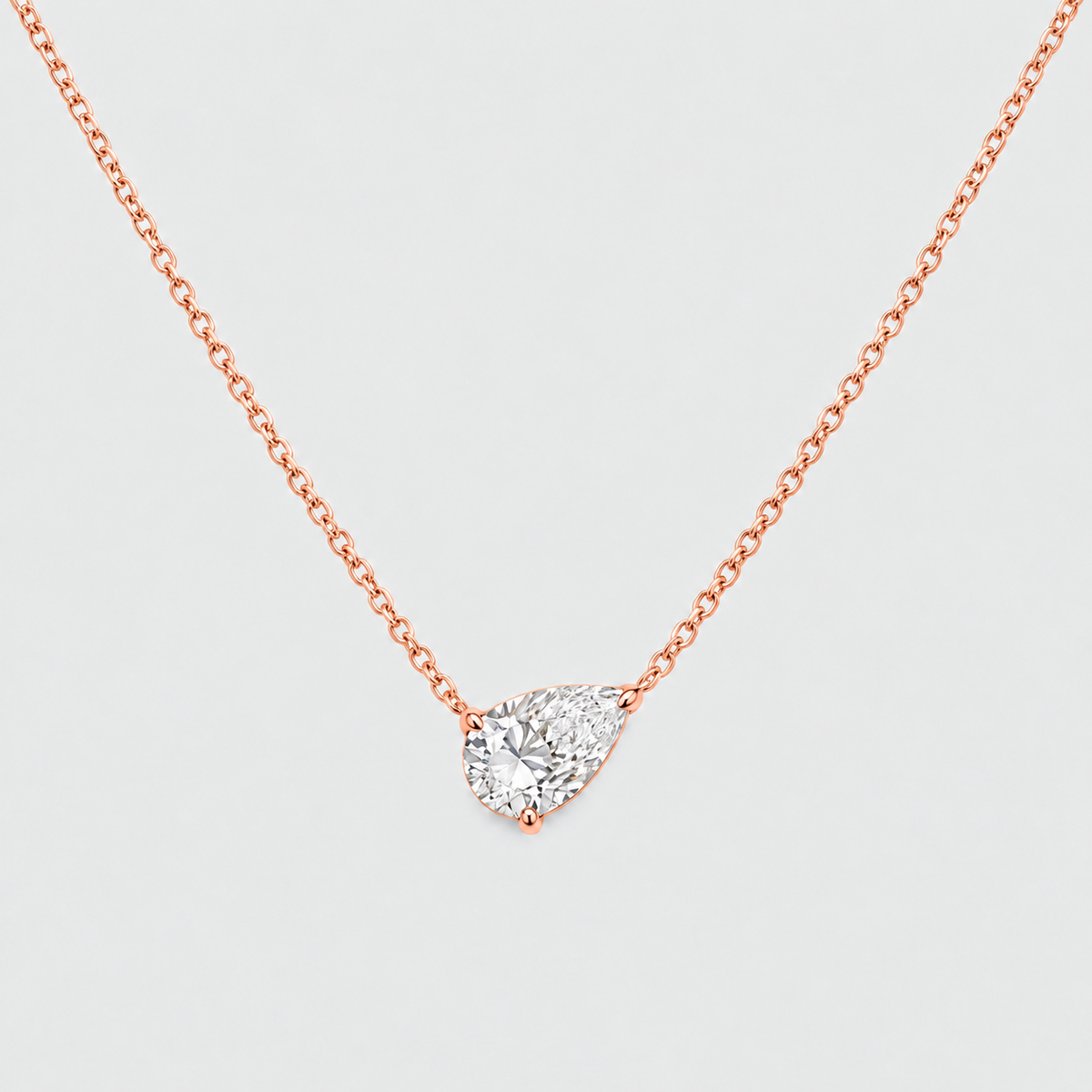 A rose gold-toned chain necklace with a pear-cut clear gemstone pendant in a prong setting, shown on a light gray background.