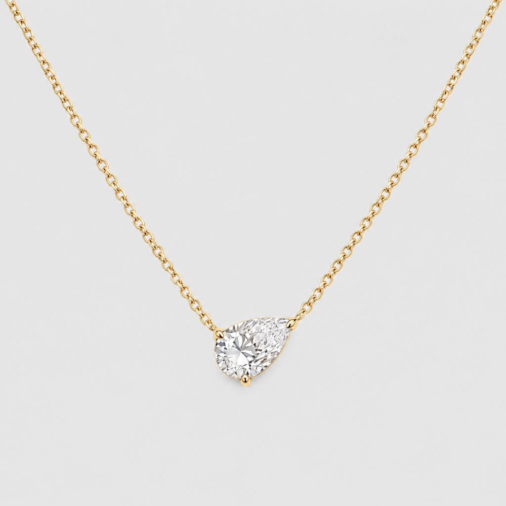 A yellow gold-toned chain necklace with a pear-cut clear gemstone pendant in a prong setting, shown on a light gray background.