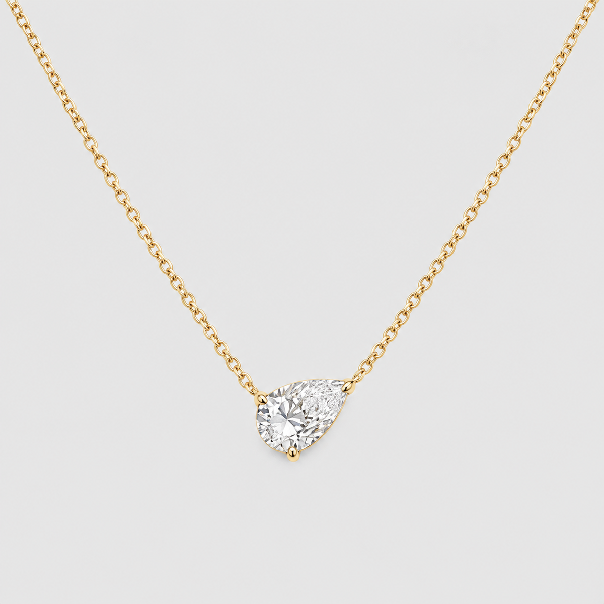 A yellow gold-toned chain necklace with a pear-cut clear gemstone pendant in a prong setting, shown on a light gray background.