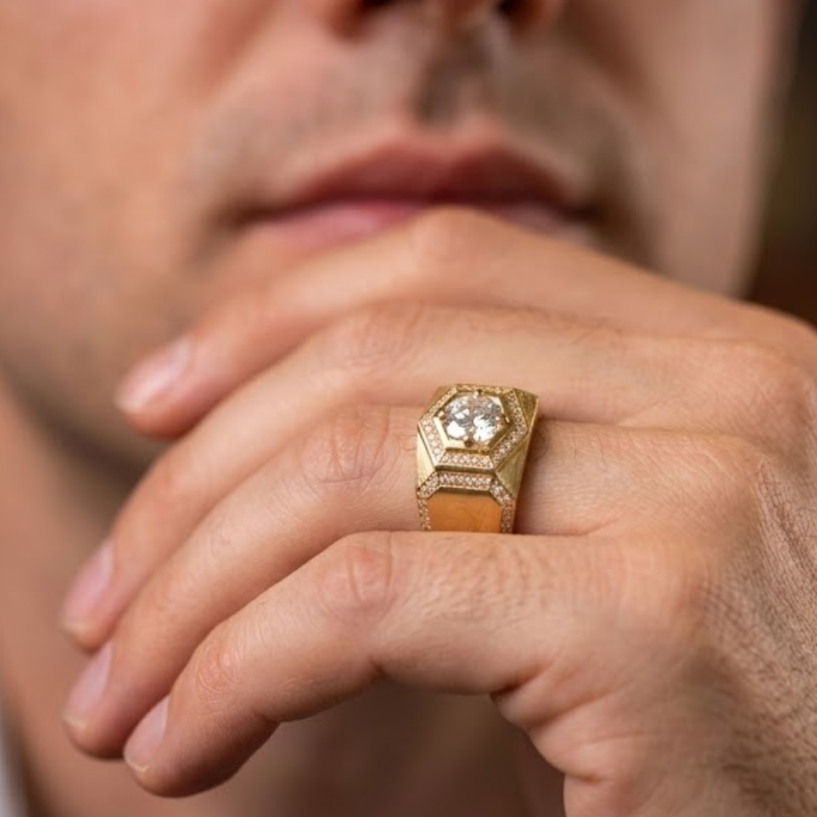 A close-up of a man's hand wearing a gold ring with a large clear gemstone on a warm brown background.