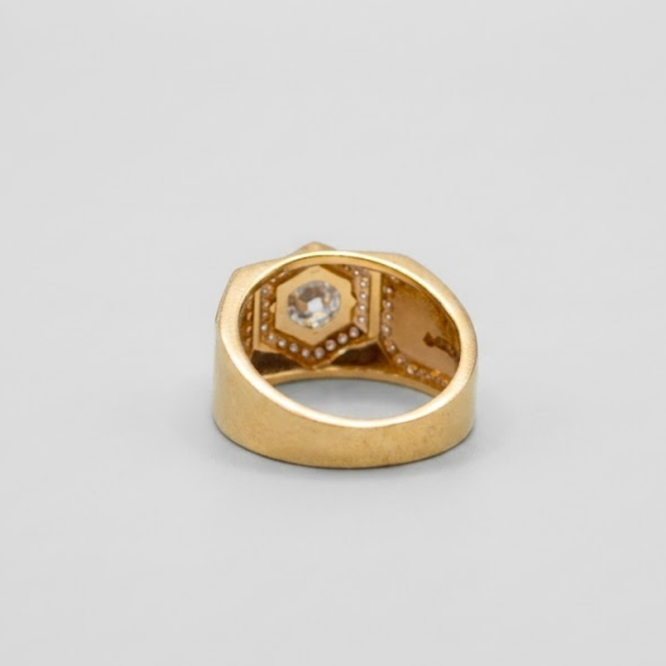 A gold ring with a geometric, multi-faceted setting and a central clear gemstone, photographed on a neutral light gray background.