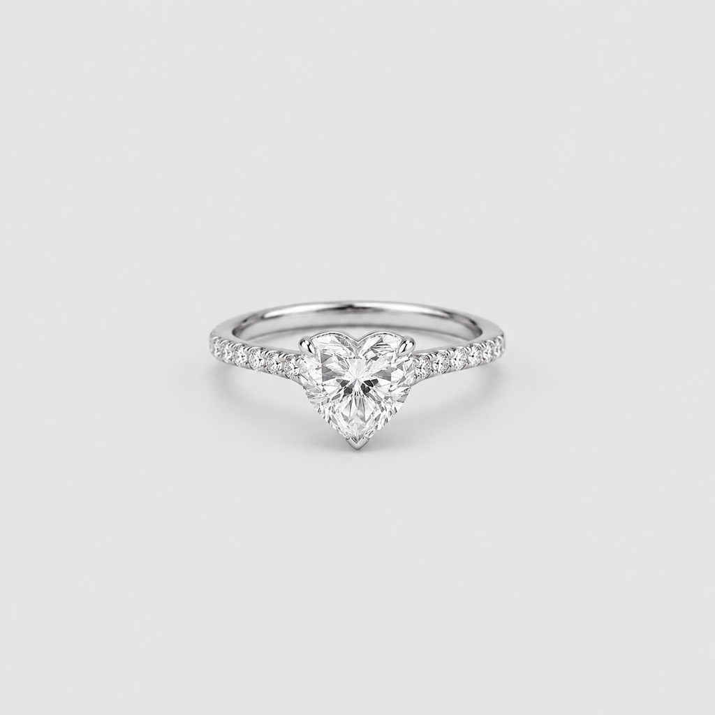 A heart-shaped diamond engagement ring with a silver-colored band set with small pavé diamonds, centered on a light gray background.