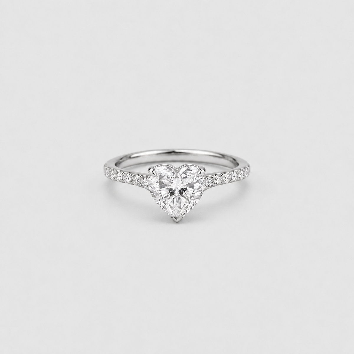 A heart-shaped diamond engagement ring with a silver-colored band set with small pavé diamonds, centered on a light gray background.