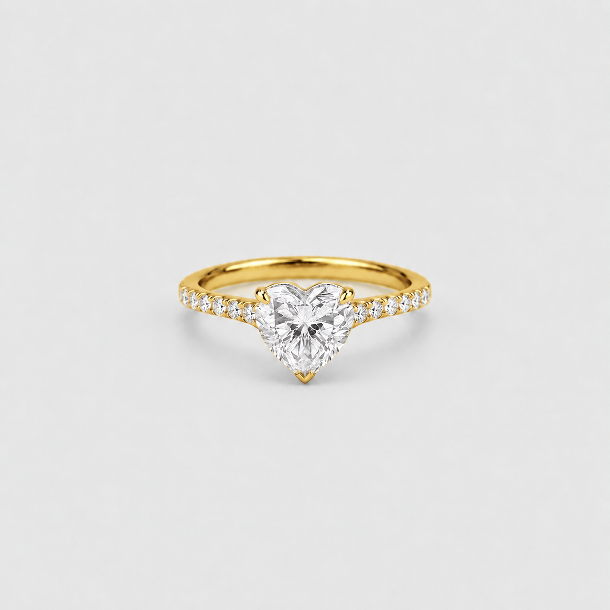 A yellow gold ring featuring a heart-shaped clear gemstone in a prong setting, with small pavé diamonds along the band, shown on a light gray background.
