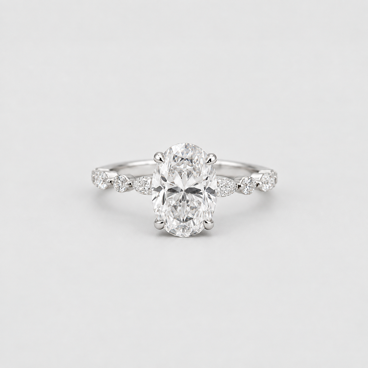 An oval-cut diamond engagement ring with a silver-toned band accented by small round diamonds, photographed on a clean light gray background.