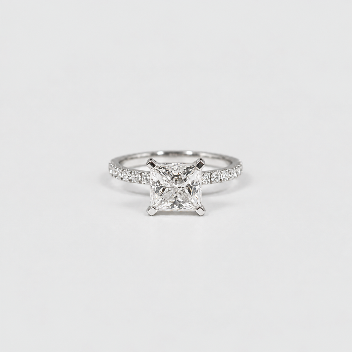 A silver engagement ring featuring a princess-cut diamond center stone and a pavé-set band, photographed on a clean white background.