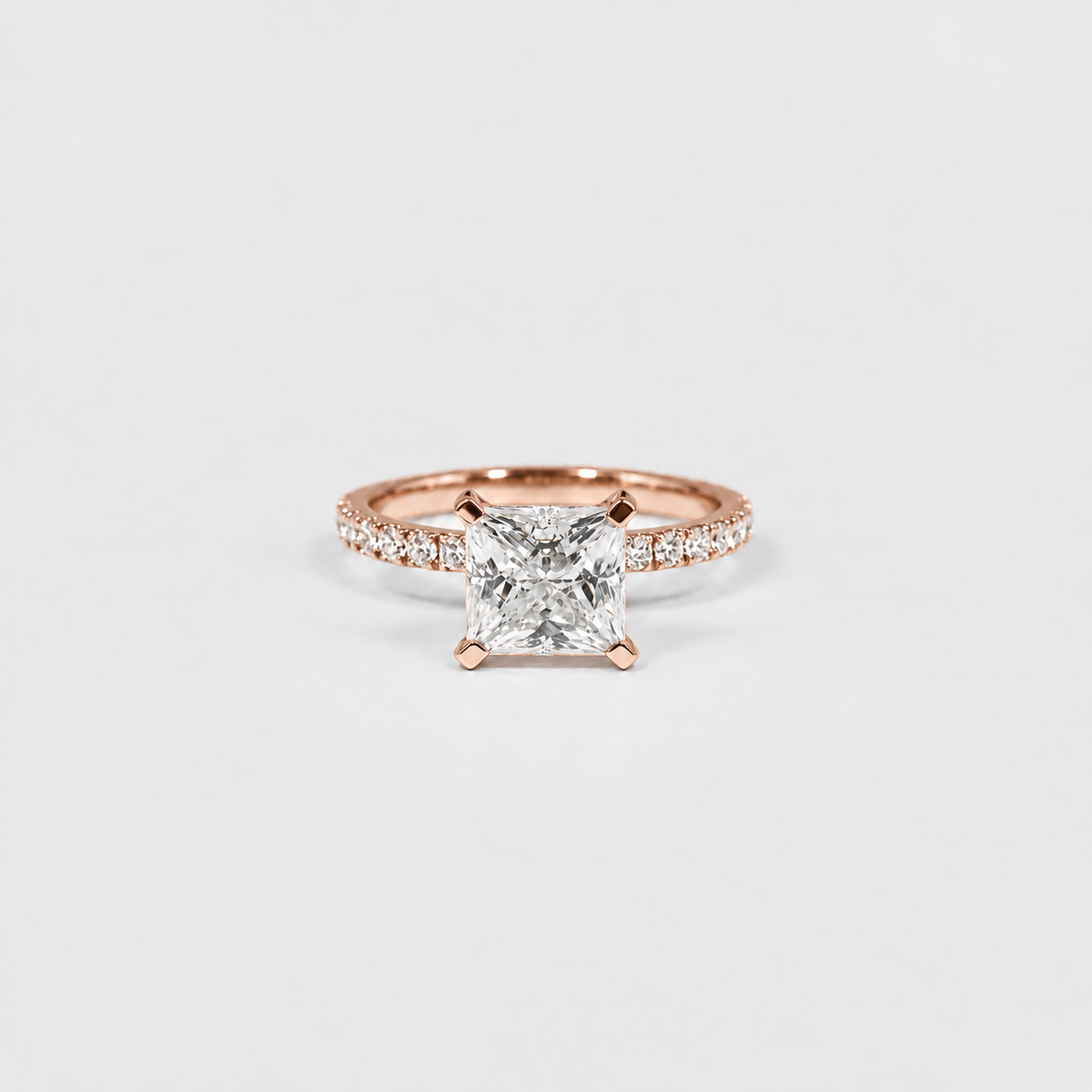 A rose gold solitaire engagement ring featuring a princess-cut diamond center stone and a pavé band, set against a clean white background.