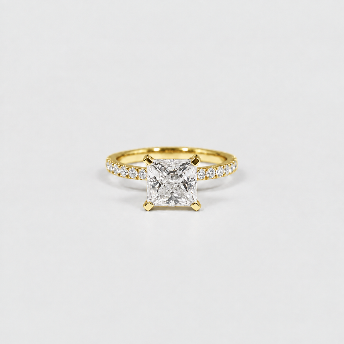 A yellow gold solitaire engagement ring featuring a princess-cut diamond center stone and a pavé diamond band, set against a clean white background.