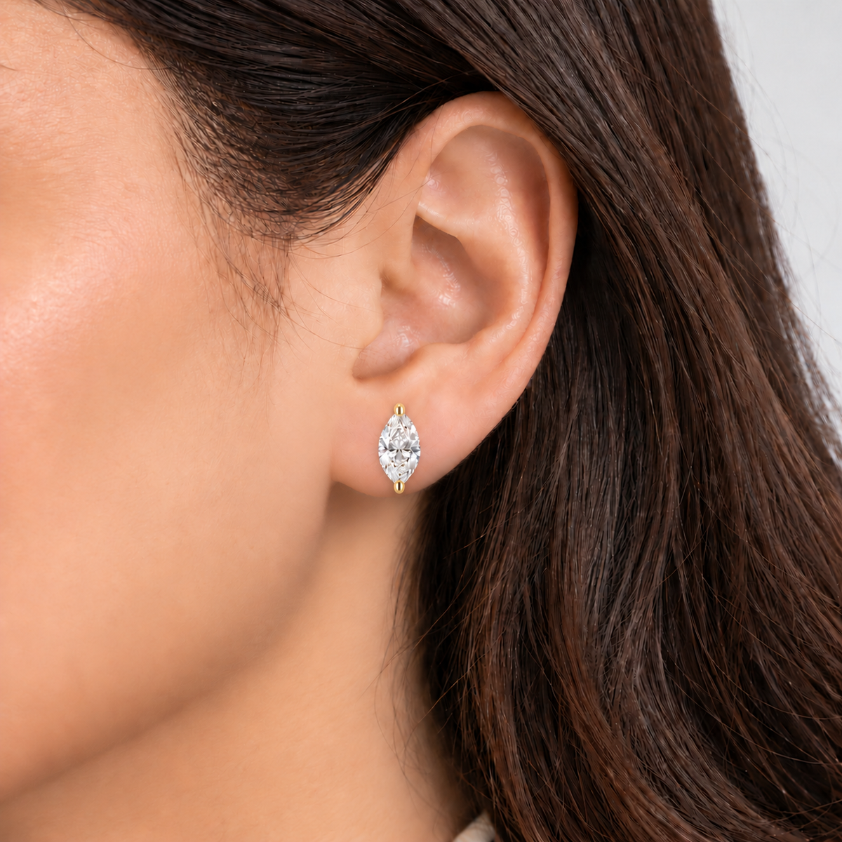 A close-up of a woman's ear wearing a small marquise-cut diamond stud earring in yellow gold.