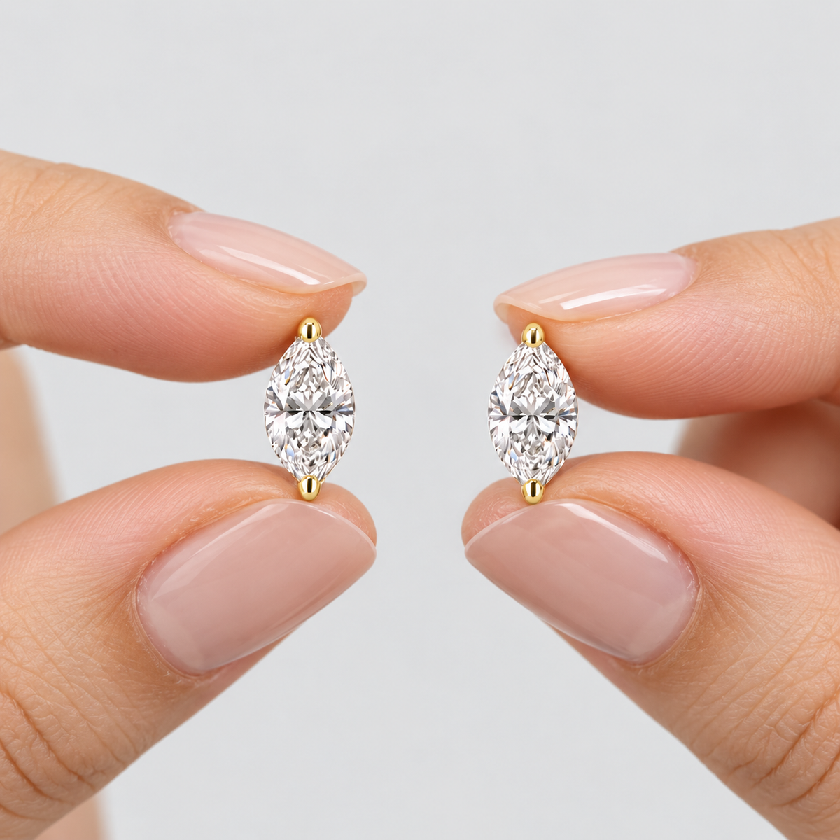 A close-up of two hands with pale pink manicured nails holding a pair of marquise-cut diamond stud earrings in yellow gold settings against a light gray background.