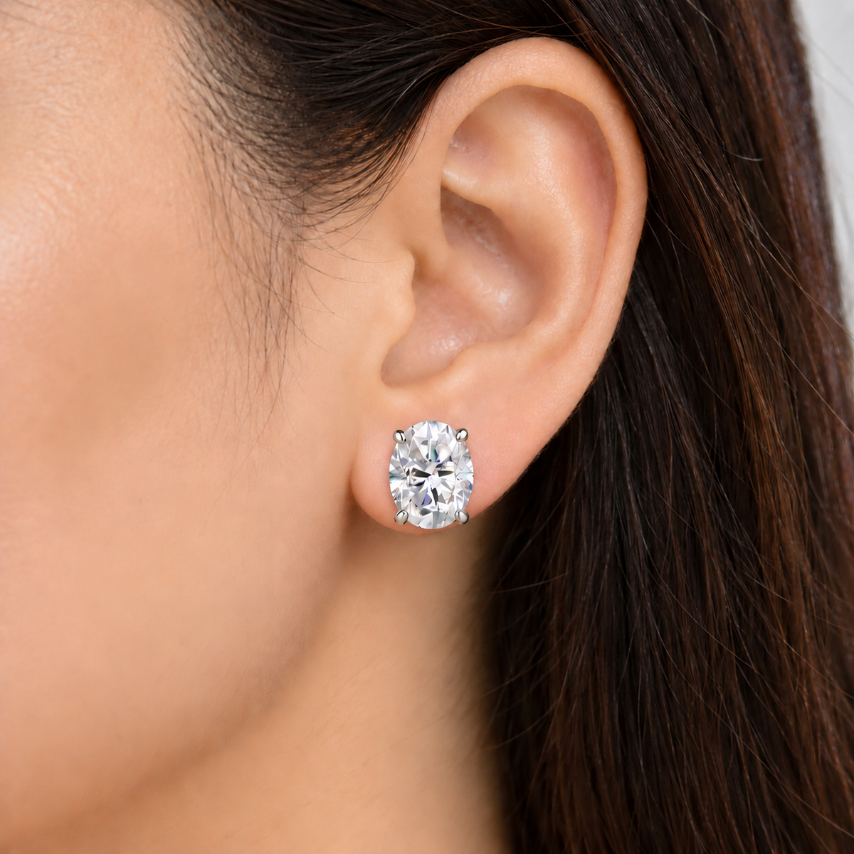 A close-up of a person's ear wearing a single oval-cut clear gemstone stud earring in a silver-toned setting.