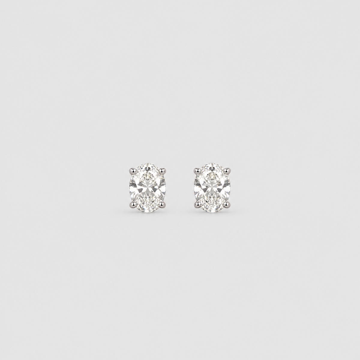 A pair of oval-cut diamond stud earrings in a silver-toned setting, centered on a light gray background.