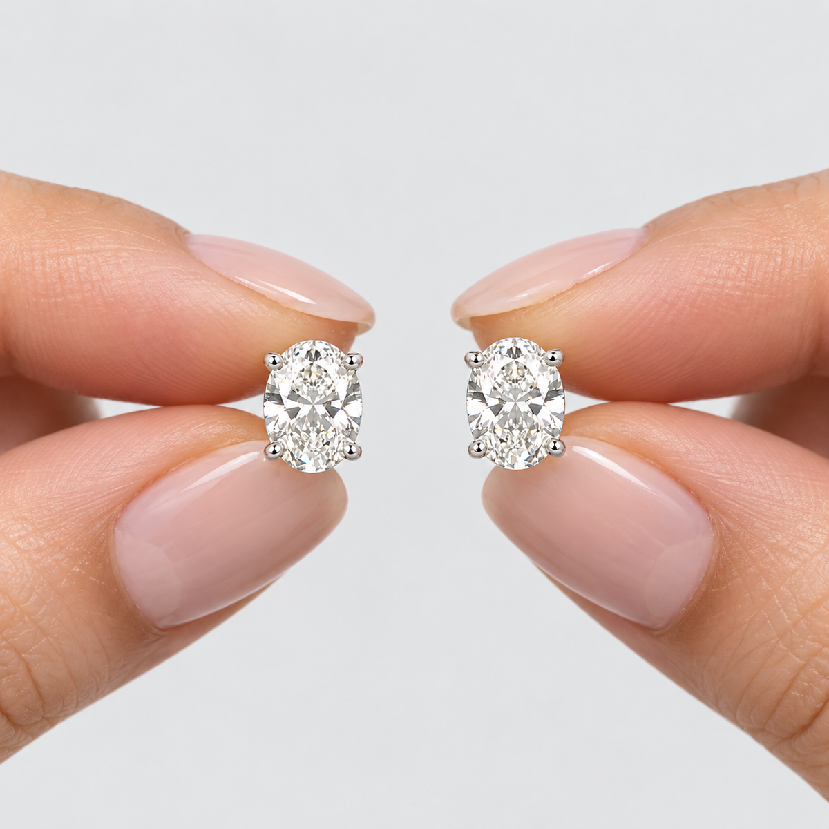 A pair of oval-cut diamond stud earrings held between two hands with neatly manicured fingernails against a clean, light gray background.