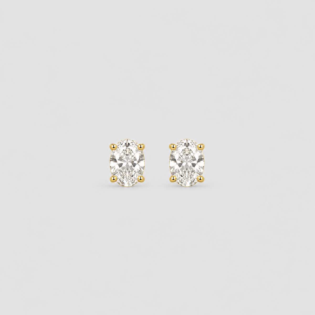 A pair of oval-cut diamond stud earrings in yellow gold prong settings, centered on a light gray background.