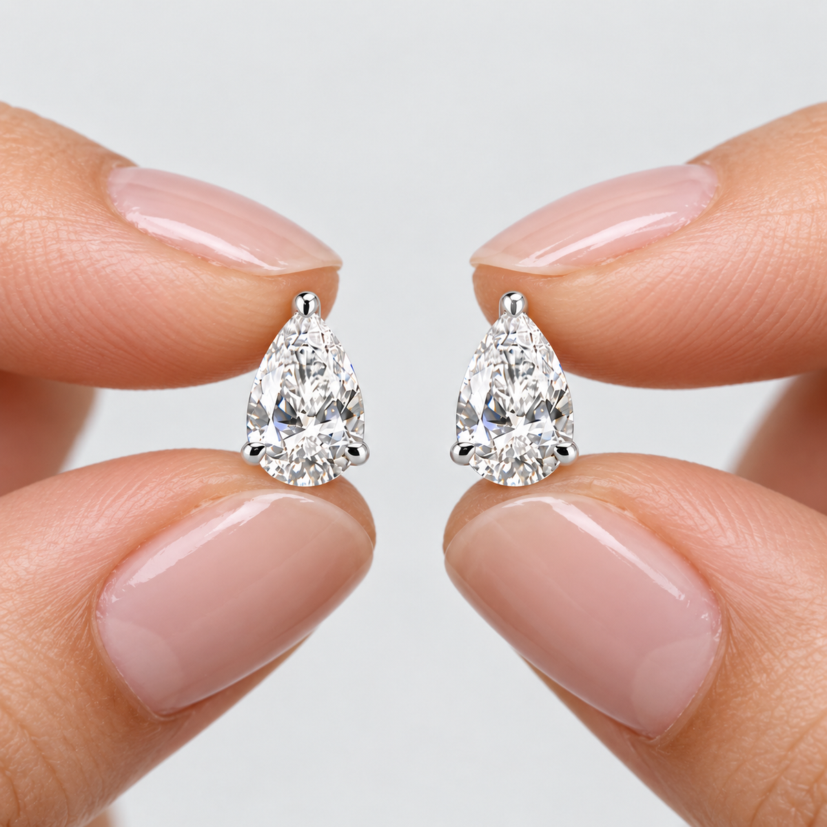A close-up of a person's fingers with light pink manicured nails holding a pair of pear-cut diamond stud earrings in a silver setting against a light gray background.