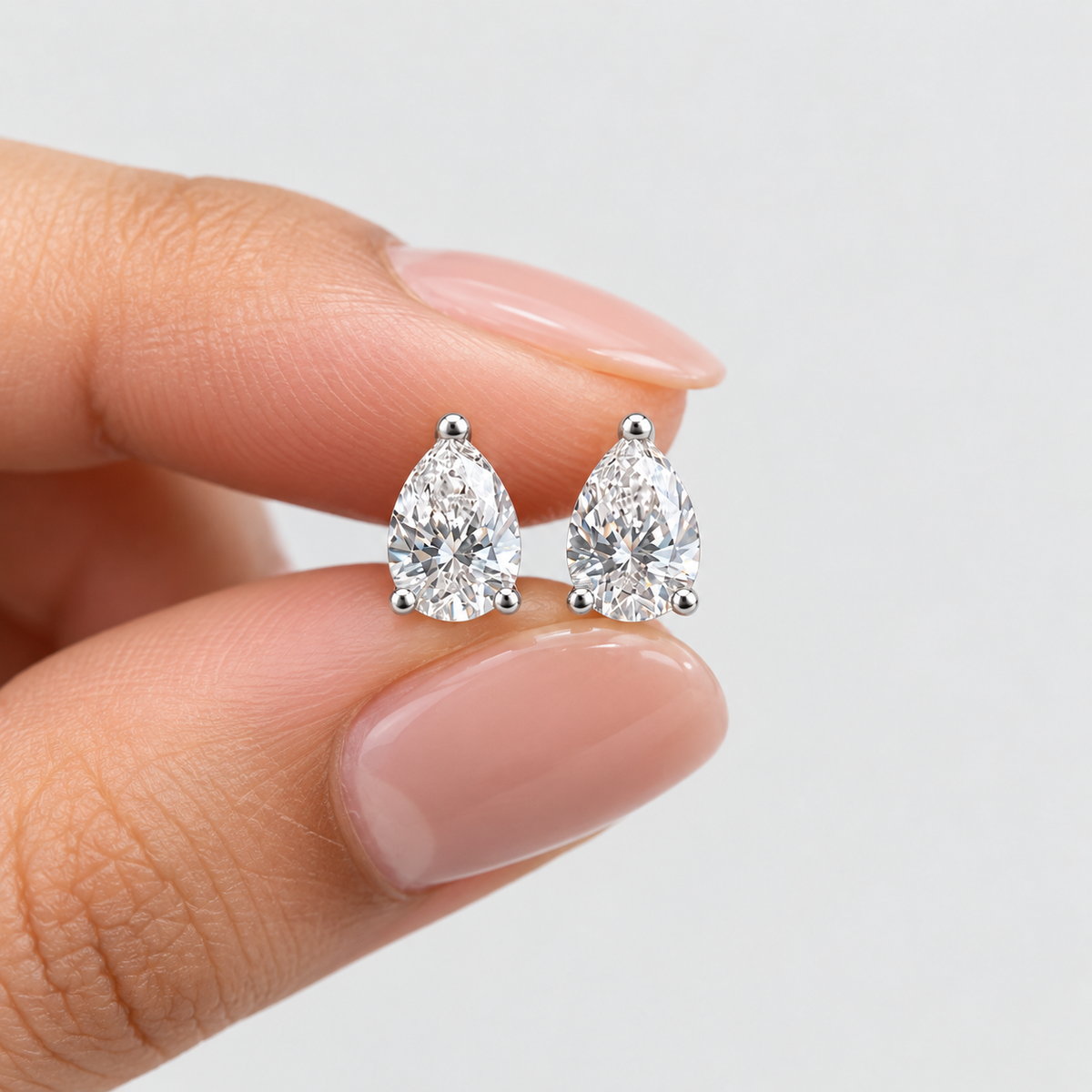A close-up of a person's hand with manicured nails holding a pair of pear-cut diamond stud earrings in silver settings against a clean white background.