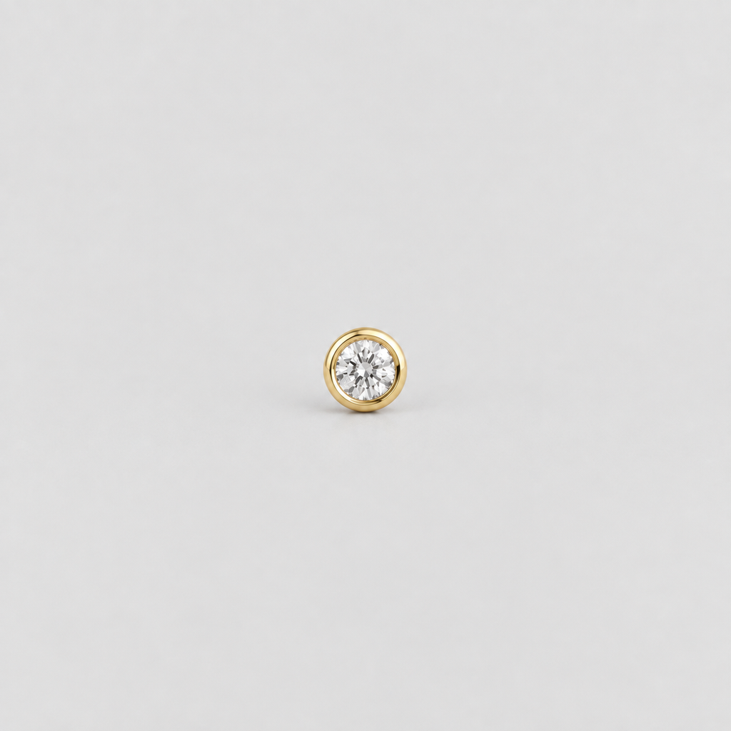 A single gold stud earring featuring a round bezel-set clear gemstone, centered on a light gray background.