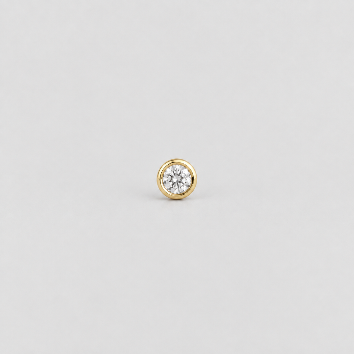 A single gold stud earring featuring a round bezel-set clear gemstone, centered on a light gray background.