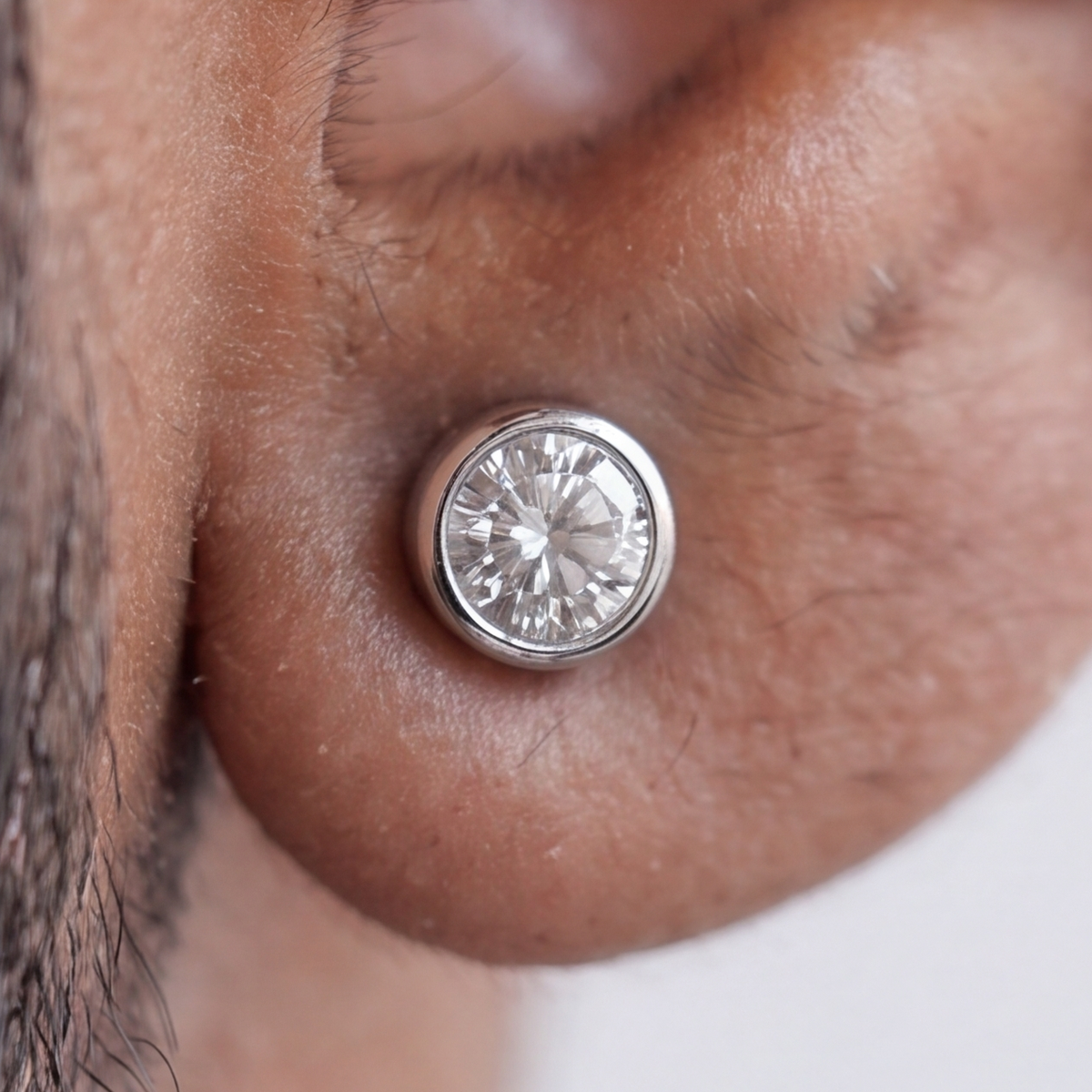 A close-up shot of a silver-toned circular stud earring with a clear round-cut gemstone, worn on a person's earlobe.