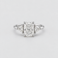 An elegant diamond ring featuring a central rectangular radiant-cut diamond flanked by two small triangular diamonds on a polished silver-colored band, set against a clean light gray background.