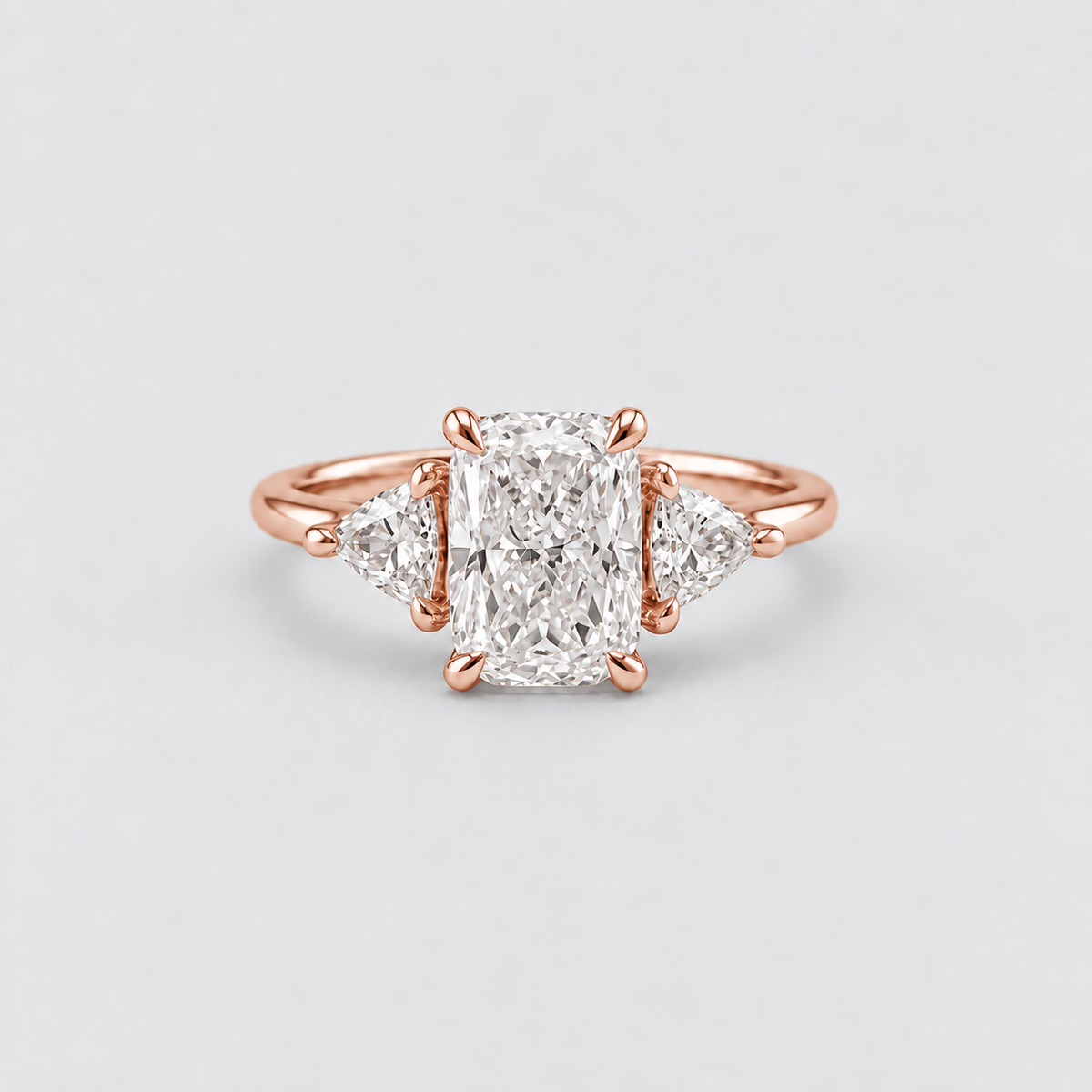 A rose gold engagement ring featuring a central rectangular radiant diamond flanked by two smaller round diamonds, set on a clean light gray background.