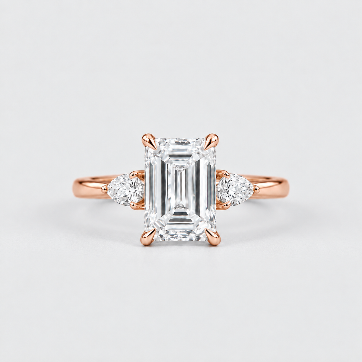 An emerald-cut diamond engagement ring in rose gold with small round diamonds on either side of the band, set against a clean light gray background.