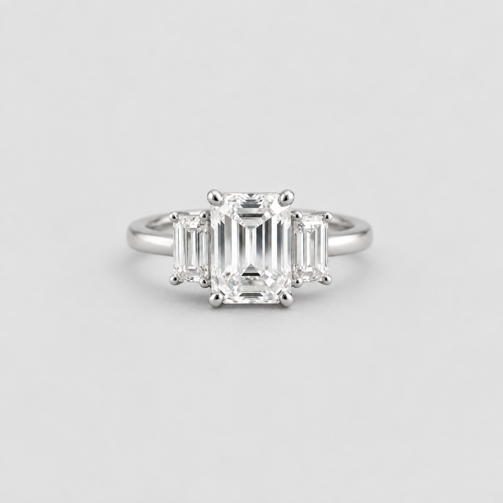 A three-stone diamond ring featuring a central emerald-cut diamond flanked by two baguette-cut diamonds, set in a polished silver-colored metal band, shown against a light gray background.