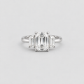 A three-stone diamond ring featuring a central emerald-cut diamond flanked by two baguette-cut diamonds, set in a polished silver-colored metal band, shown against a light gray background.
