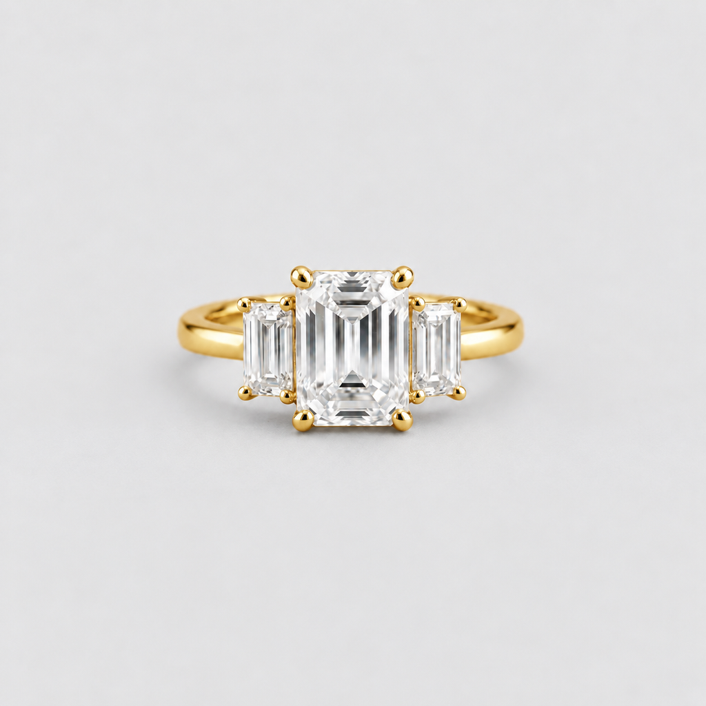 A yellow gold three-stone ring featuring a large emerald-cut center diamond flanked by two smaller baguette-cut diamonds, set against a clean light gray background.