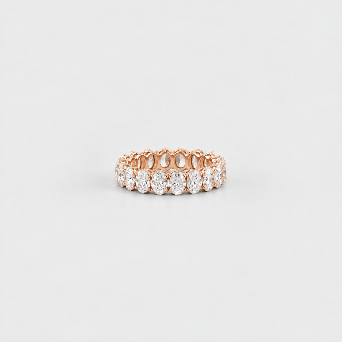 A rose gold eternity ring featuring a row of round-cut clear gemstones, photographed on a clean, light gray background.