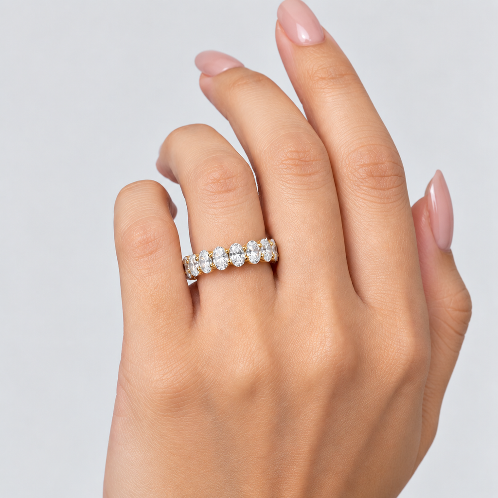 A close-up of a hand wearing a silver eternity ring set with round-cut clear gemstones, shown against a light gray background.