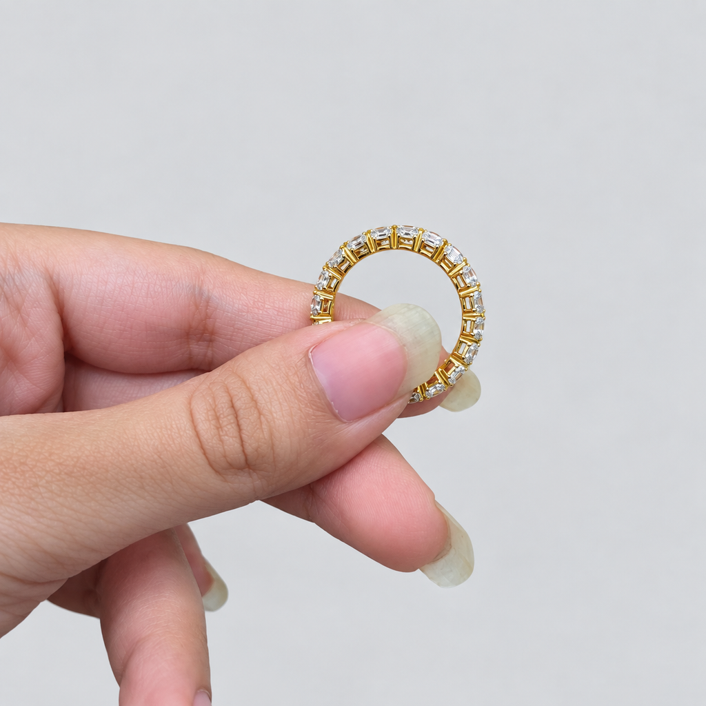 A close-up of a hand holding a gold ring with small clear stones set around the band, against a plain light gray background.
