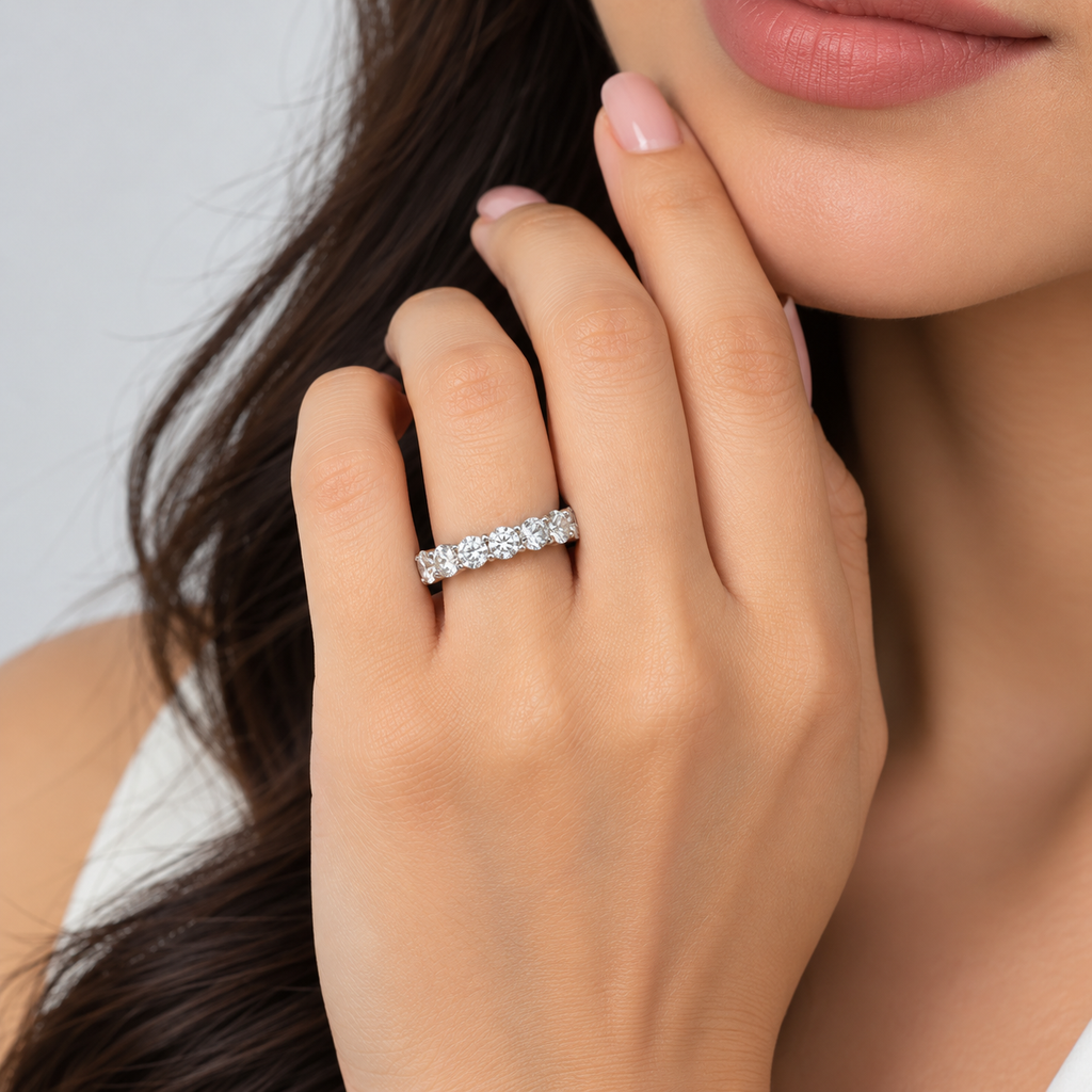 A close-up lifestyle shot of a woman's hand wearing a five-stone diamond eternity ring. The hand is resting near her chin, showing her skin, nails, and dark hair against a soft, light gray background.