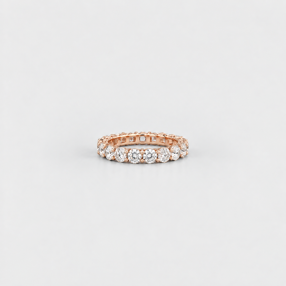 A rose gold eternity ring featuring a row of round-cut clear gemstones, photographed on a clean, light gray background.