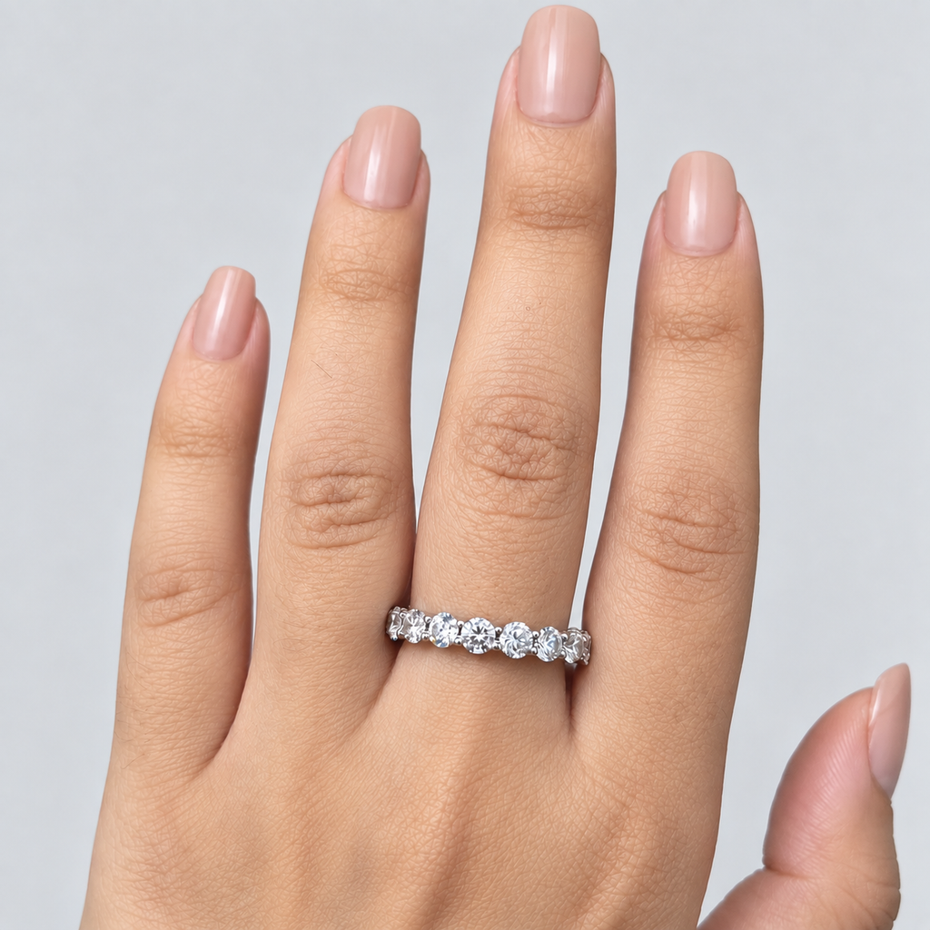 A close-up of a hand wearing a silver eternity ring set with round-cut clear gemstones, shown against a neutral light gray background.