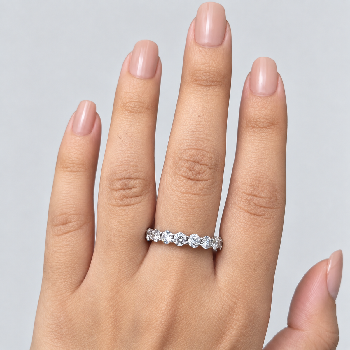 A close-up of a hand wearing a silver eternity ring set with round-cut clear gemstones, shown against a neutral light gray background.