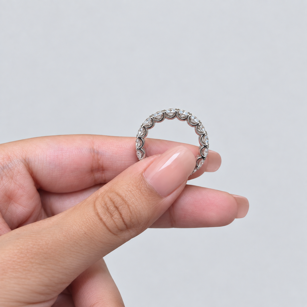 A close-up of a hand holding a thin silver band ring with a row of small clear stones around the circumference, set against a plain light gray background.