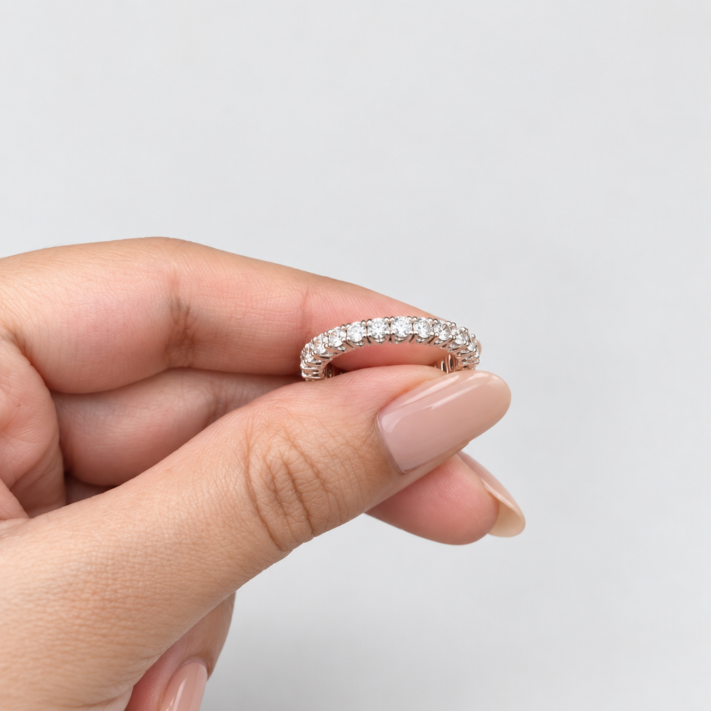 A close-up of a hand holding a thin diamond eternity ring with a silver-colored setting against a plain light gray background.