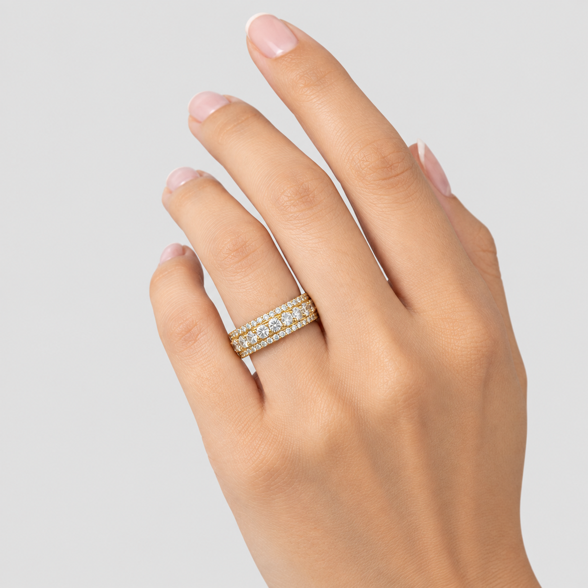 A close-up of a hand wearing a delicate double-row diamond band ring in yellow gold, set against a clean light gray background.