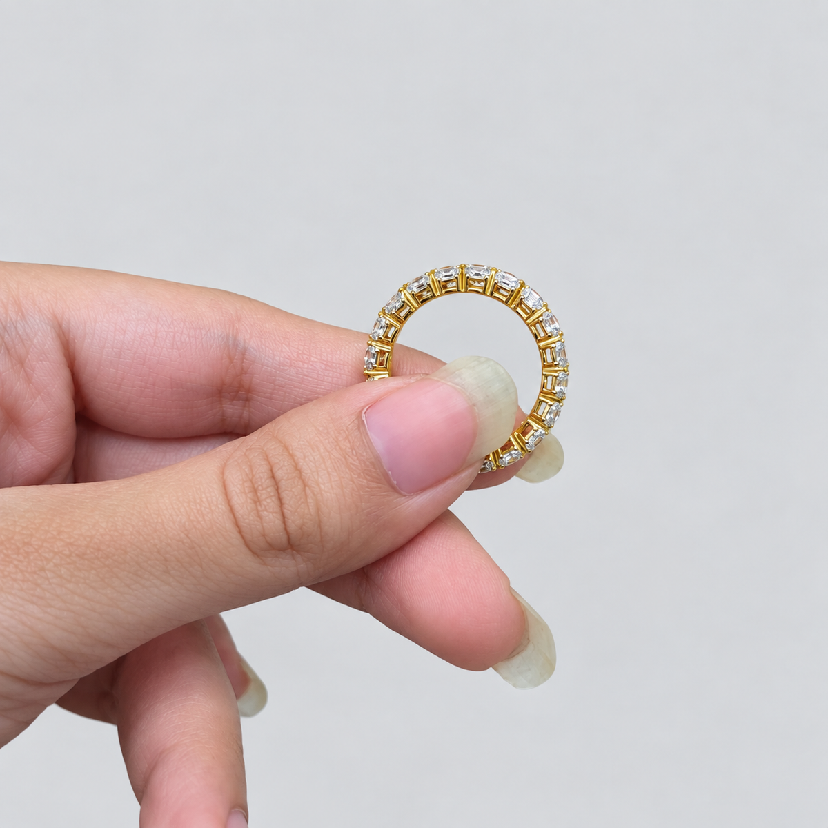 A close-up of a hand holding a gold ring with small clear stones set around the band, against a plain light gray background.