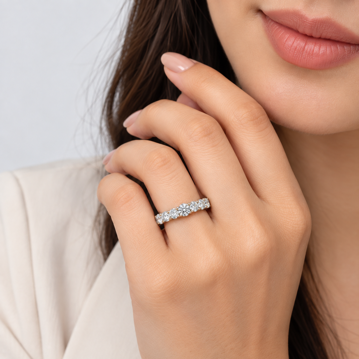 A close-up lifestyle shot of a woman wearing a silver diamond eternity ring on her finger, with her hand resting near her face against a soft light gray background.