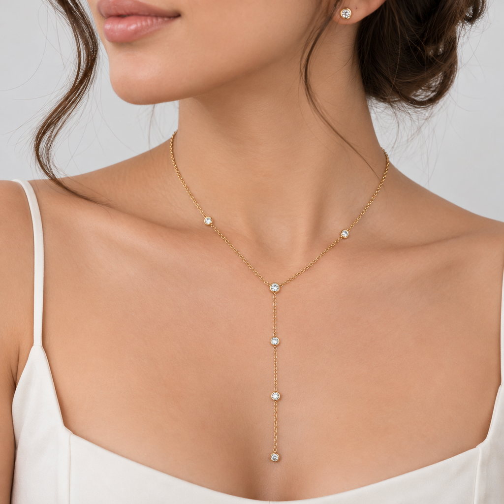 A close-up of a woman wearing a delicate gold Y-necklace with five small clear gemstones, paired with a matching stud earring, set against a clean light gray background.