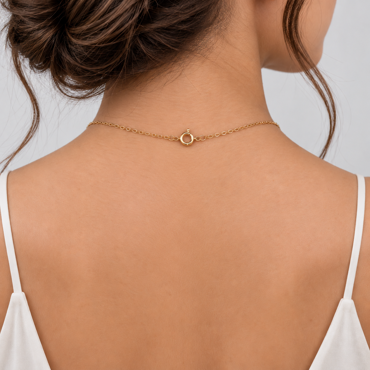 A close-up back view of a woman wearing a delicate gold chain necklace with a small circular ring charm, styled with a white spaghetti strap top against a light gray background.