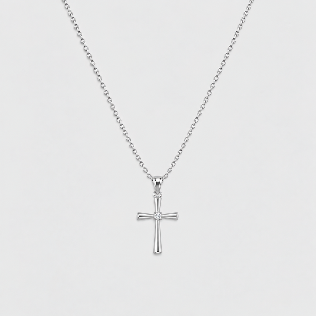 A silver cross pendant necklace with a small clear gemstone at the center, suspended from a delicate silver ball chain against a light gray background.