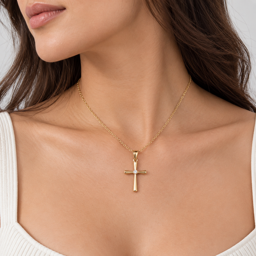 A gold cross pendant necklace worn by a model, featuring a small white crystal accent on the cross, set against a clean white ribbed top.