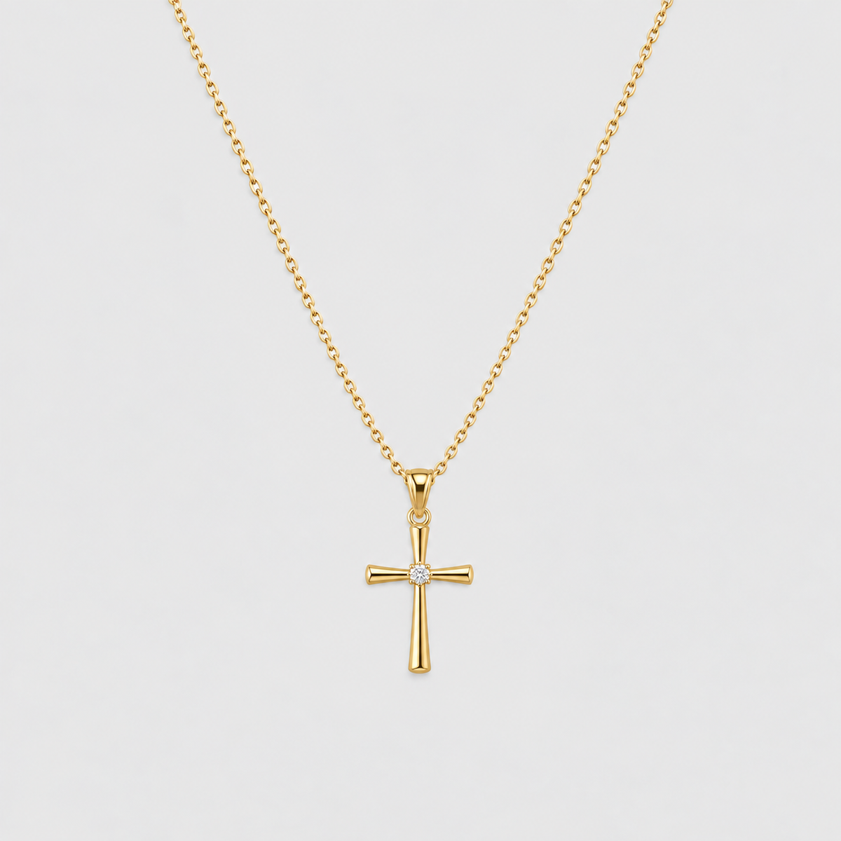 A gold cross pendant necklace with a small clear gemstone at the center, suspended from a delicate gold chain against a light gray background.