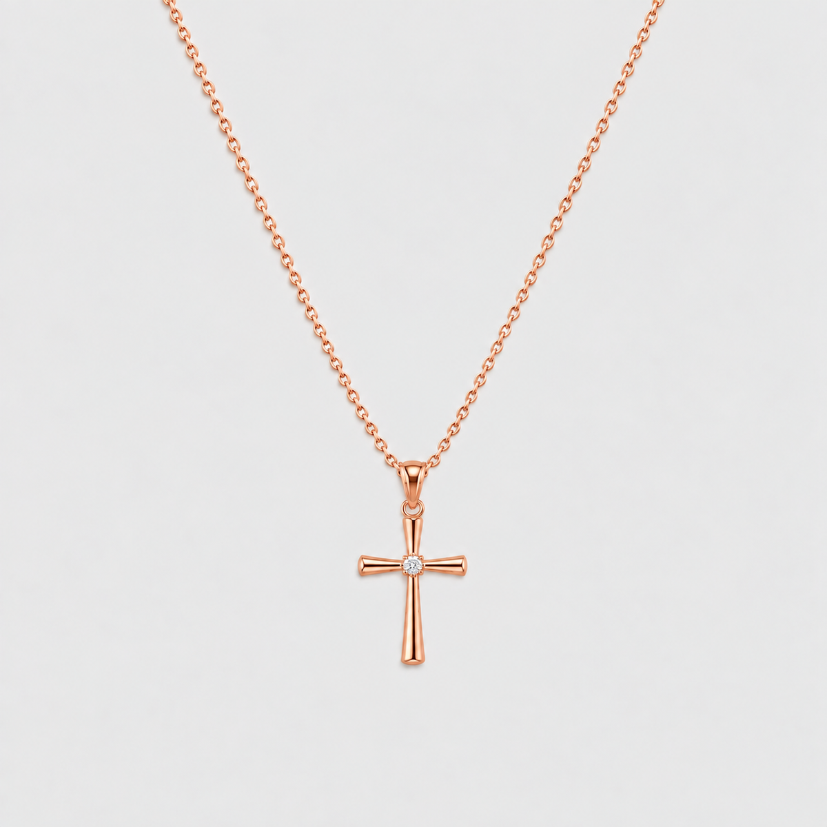 A rose gold cross pendant necklace with a small clear gemstone at the center, suspended from a delicate matching chain against a light gray background.