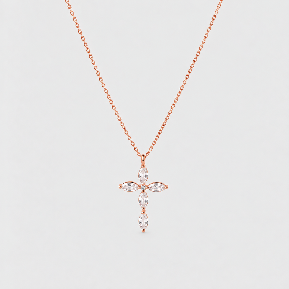 A delicate rose gold chain necklace featuring a cross-shaped pendant adorned with marquise-cut clear gemstones, set against a solid light gray background.