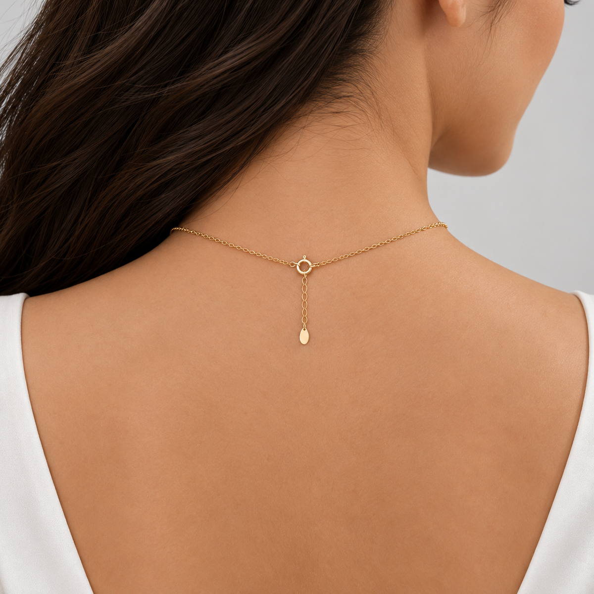 A close-up back view of a woman wearing a delicate gold chain necklace with a small circular ring detail and a dangling teardrop charm, styled with a white top against a light gray background.